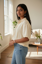 Woman wearing a beige blouse and blue jeans standing in a bright room with large windows.