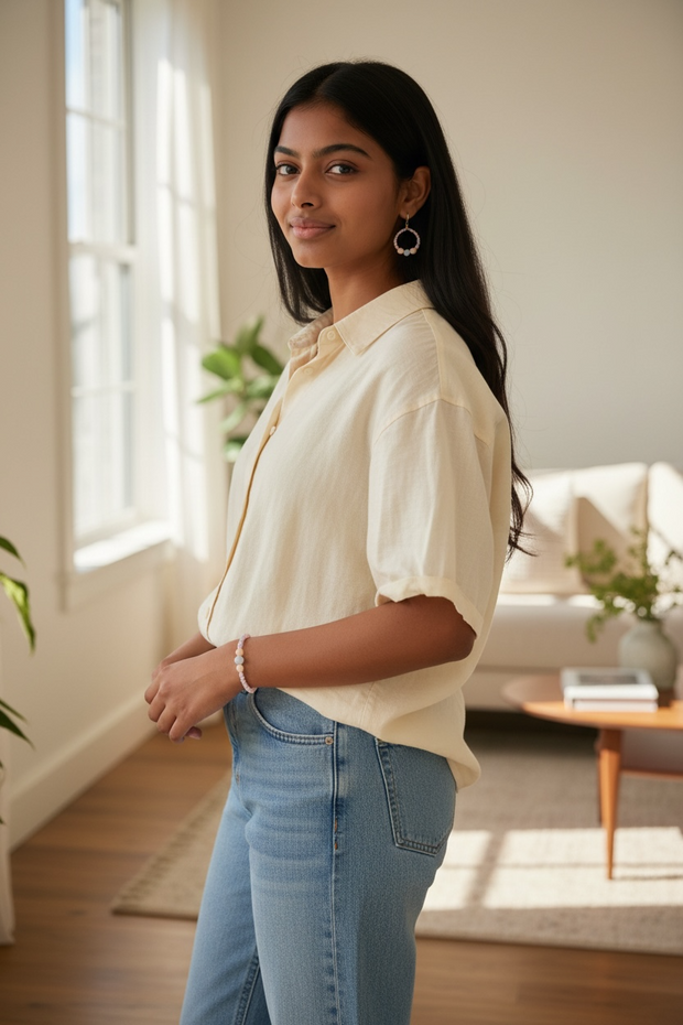 Woman wearing a beige blouse and blue jeans standing in a bright room with large windows.